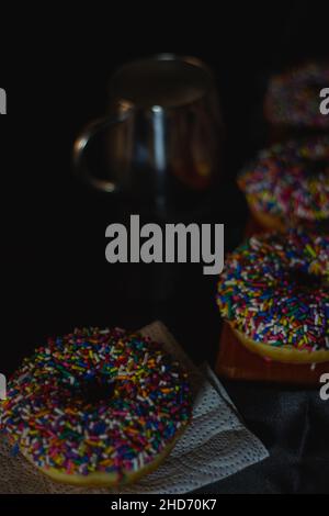 Vertical shot of three delicious donuts with sprinkles on a dark background with a mug Stock Photo