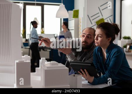 Architect pointing pen at skyscraper architectural model collaborating with colleague engineer holding tablet with blueprints in modern office. Architects looking inspired collaborating on maquette. Stock Photo