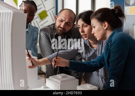 Two women professional architects explaining construction model design to team colleagues in front of white foam maquette. Engineers working together on residential project in architectural office. Stock Photo