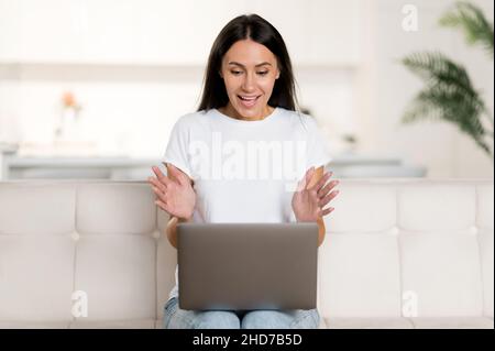 Caucasian young adult beautiful happy brunette woman sitting on sofa in living room using laptop, talking via video conference, gesturing with hands, looking at laptop screen, smiling friendly Stock Photo