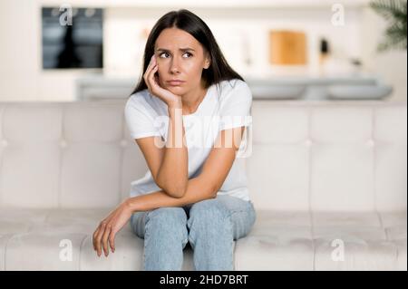 Disappointed unhappy caucasian young woman, sitting on the sofa in the living room, she has personal problems, failure at work, depression, bad mood, sad girl is lonely at home Stock Photo