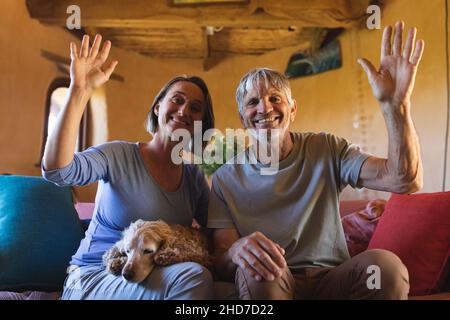 Portrait of mature couple with dog waving hands while sitting on couch in cob home Stock Photo
