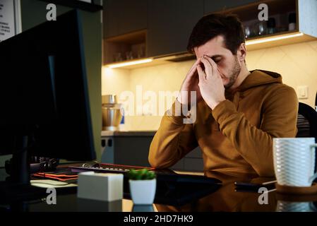 Man sitting at table at home office and works with computer. Freelancer remotely working late. Emotional stress and burnout Stock Photo