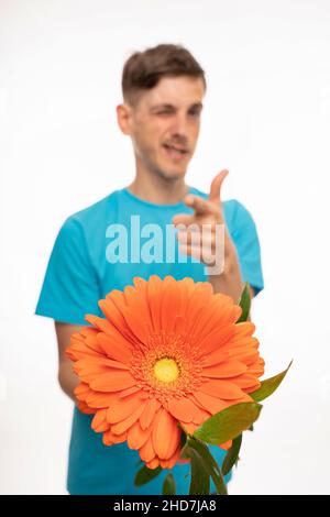 Young handsome tall slim white man with brown hair offering orange flower winking with one eye with blue shirt isolated on white background Stock Photo