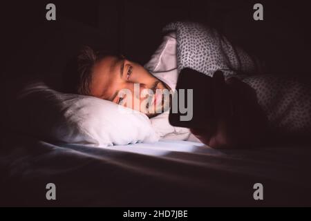 Young man lying in bed and using smartphone at night before sleeping. Social media addiction and insomnia. Communication and people concept  Stock Photo
