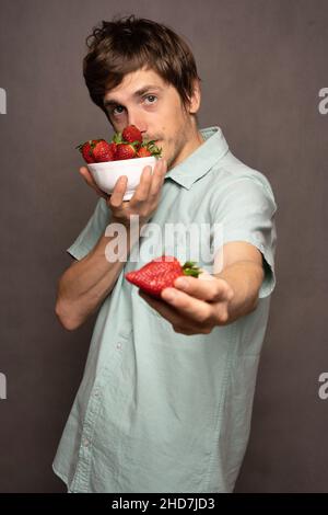 Young handsome tall slim white man with brown hair sniffing strawberries holding one in light blue shirt on grey background Stock Photo