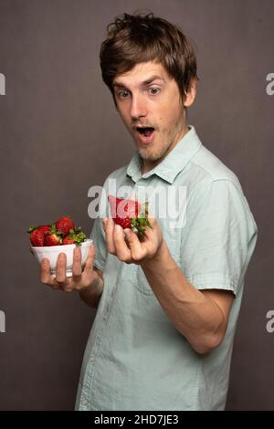 Young handsome tall slim white man with brown hair shocked by a strawberry in light blue shirt on grey background Stock Photo