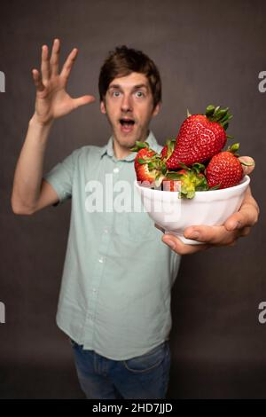 Young handsome tall slim white man with brown hair holding bunch of strawberries shocked in light blue shirt on grey background Stock Photo