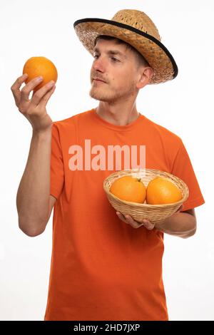 Young handsome tall slim white man with brown hair observing an orange closely in orange shirt with straw hat isolated on white background Stock Photo