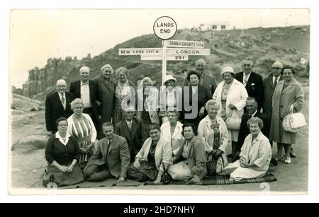 Original postcard of happy pensioners, men and women, wearing suits, carrying handbags, on an excursion in late Summer, probably coach tour group, to Land's End. They pose by the famous signpost, showing John O'Groats, London and New York mileage, printed on reverse Richards Bros. 3 Sept 1962, Cornwall, U.K. Stock Photo