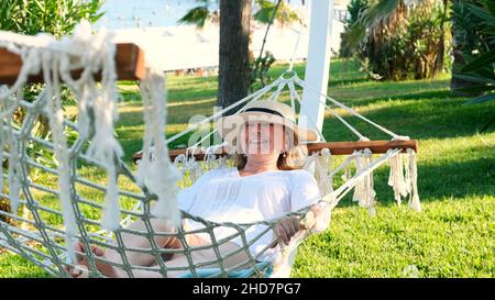 elderly woman in a straw hat is resting in a hammock around the palm trees and enjoying the tranquility and relaxation. Senior citizen lifestyle Stock Photo