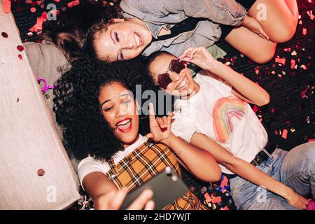 Making memories with crazy selfies. Overhead view of three happy friends taking a selfie while lying on the floor at a house party. Group of cheerful Stock Photo