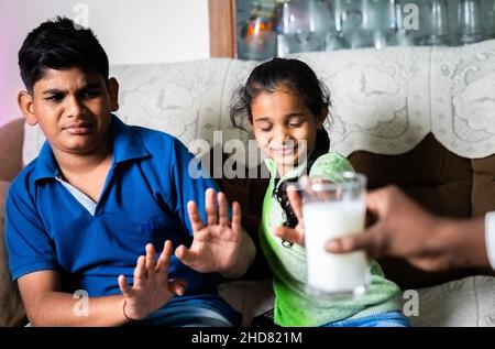 Indian teenager sibling kids saying no or refusing for drinking milk to parents while watching television at home - concept of unhealthy food habits Stock Photo
