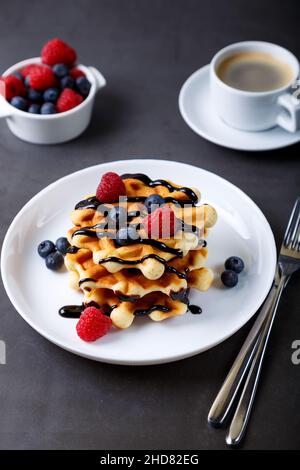 Viennese or Belgian waffles with fresh berries (raspberries and blueberries) on a white plate and a cup of coffee. Traditional dessert. Close-up. Stock Photo