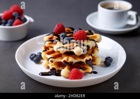 Viennese or Belgian waffles with fresh berries (raspberries and blueberries) on a white plate and a cup of coffee. Traditional dessert. Close-up. Stock Photo