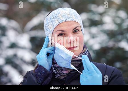 Young woman in warm winter clothes, wearing protection gloves, putting or removing disposable single use blue face virus mouth mask, closeup, blurred Stock Photo
