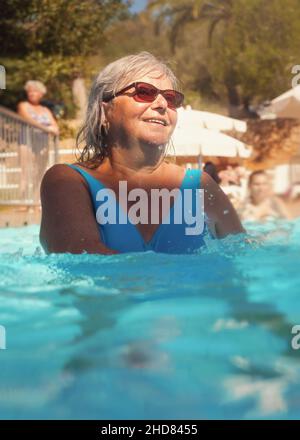 Elderly senior woman with grey hair, wearing blue swimsuit doing water aerobics in hotel pool Stock Photo