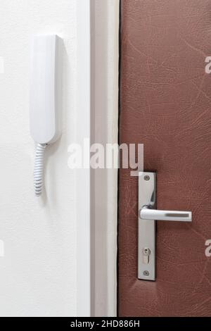 Simple white intercom or house phone attached to a wall next to a closed door with metallic handle. Home safety and communication. Stock Photo