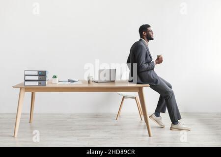 Handsome young black businessman leaning on table with cup of coffee, taking break from online job at home office Stock Photo