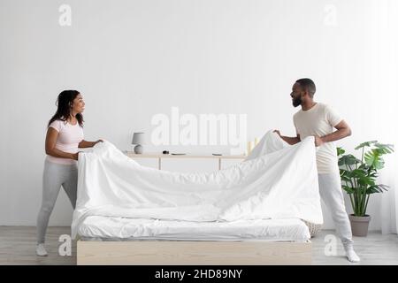 Cheerful young black woman and guy make bed and hold blanket in bedroom interior Stock Photo