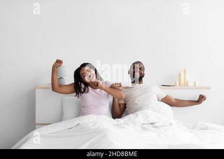 Cheerful young black boyfriend and girlfriend wake up after night sleep and stretch body on bed Stock Photo