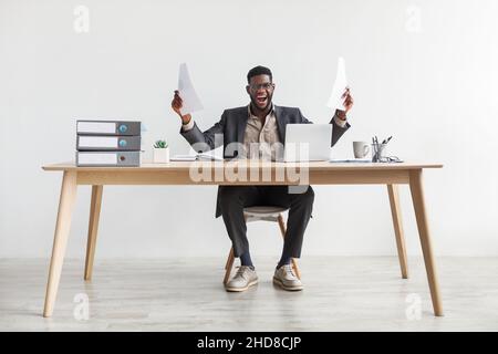 Emotional young black man with documents sitting at desk in front of laptop, feeling triumphant, shouting in excitement Stock Photo