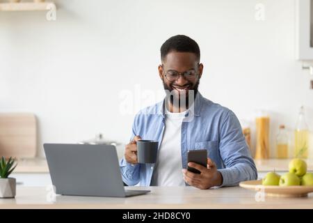 Smiling young black male with beard in glasses drinks coffee from cup, checks app on smartphone Stock Photo