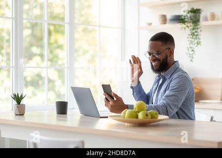 Friendly smiling millennial black man with beard in glasses waving hand at phone and work on laptop Stock Photo