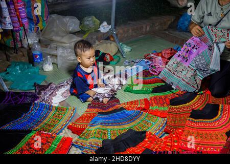Cute local chubby baby boy sits in a stall selling fabrics in the walking street night market in central Luang Prabang, northern Laos, south-east Asia Stock Photo