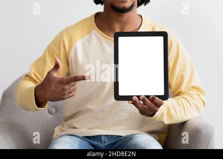 Black Guy Pointing At Digital Tablet With Blank Screen, Sitting In Armchair Stock Photo