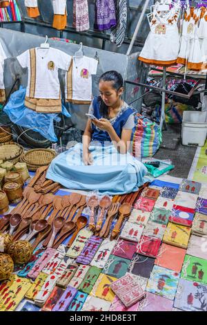 Pretty young girl stallholder in a souvenir stall looking at her mobile phone in the Walking street night market, central Luang Prabang, northern Laos Stock Photo