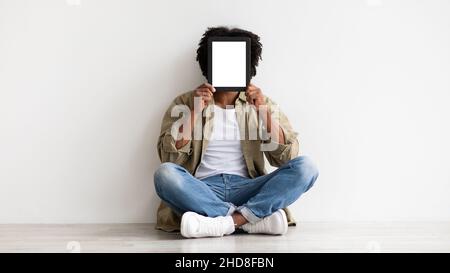 Black Man Covering Face With Blank Digital Tablet While Sitting On Floor Stock Photo