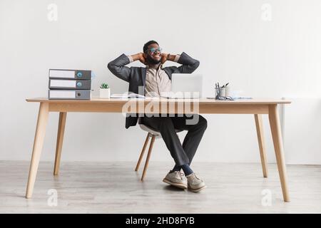Cheerful young black man relaxing on chair, sitting at table, resting with hands behind head, using laptop, taking break Stock Photo