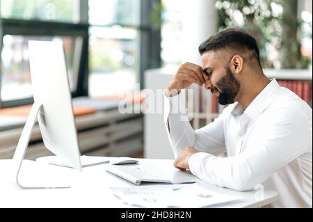 Indian young adult man upset worn out company owner, manager or office worker, sitting at his desk with closed eyes, rubbing his eyes, feeling stressed and tired from overtime, needs rest Stock Photo
