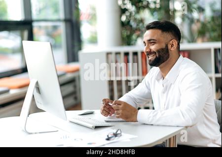 Happy successful confident young adult Indian business leader, manager or office worker with beard sitting in office using computer, video communication with colleagues or client, smiling Stock Photo