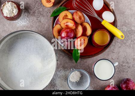 Baking process ingredients eggs, whisk, plum and baking pan on kitchen surface. Stock Photo