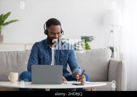 Smiling busy young black businessman freelancer or student with beard in headphones with pc making notes, study Stock Photo