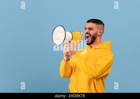 Emotional middle eastern man shouting with megaphone, making announcement or sharing news, blue background Stock Photo