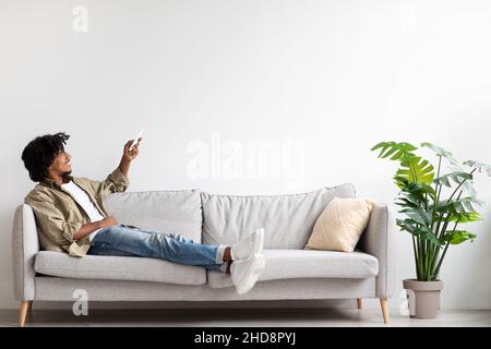 Home Air Conditioning. Black Guy Using Remote Controller While Relaxing On Couch Stock Photo