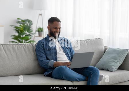 Happy young black man manager with beard sits on couch typing on pc in living room interior Stock Photo