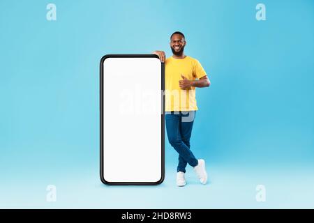 Handsome black guy standing near big cellphone with empty screen, showing thumb up, recommending cool website, mockup Stock Photo