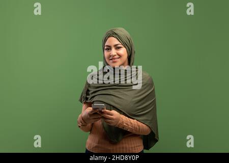 Cheerful muslim lady in hijab holding smartphone and smiling at camera, standing over dark green studio background Stock Photo