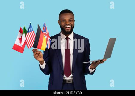 Business Language Courses. Black Businessman Holding Bunch Of Flags And Laptop Stock Photo