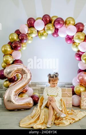 Fashionable baby girl in a white clothing celebrating her second birthday posing for camera and looking down. Lovely baby girl birthday photoshoot Stock Photo