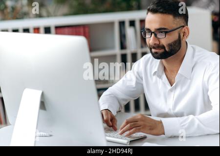 Successful Indian businessman, manager or office worker with beard and glasses, sitting at desk in modern office, wearing white shirt, using computer, texting with colleagues or client, analyzing Stock Photo