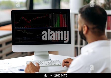 Successful smart concentrated confident Indian broke, trade analyst, sitting at computer in modern office, buying and selling cryptocurrencies, analyzing cryptocurrency market, looking at screen Stock Photo