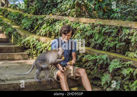 Selfie with monkeys. Young man uses a selfie stick to take a photo or video blog with cute funny monkey. Travel selfie with wildlife in Bali Stock Photo