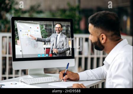 Online business video conference. Concentrated top manager or business owner sitting at desk in office, wearing white shirt, listening online business training, watching coach video, taking notes Stock Photo