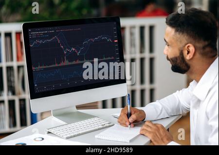 Stock trading. Focused successful Indian trader expert, stock broker, using computer in his modern office, analyzing cryptocurrency market, taking notes, planning stock strategy Stock Photo