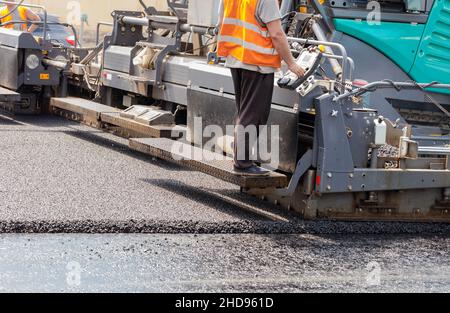 A road worker in an orange vest drives an asphalt paver and pours hot fresh asphalt onto a bitumen pavement on a summer day. Close-up, copy space. Stock Photo